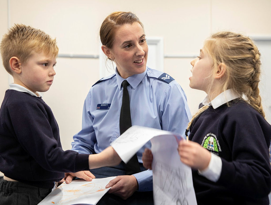 Image shows RAF aviator with school children and their drawings.